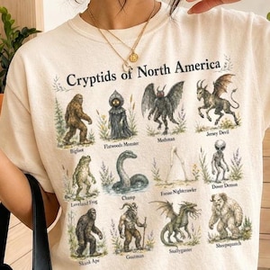 Puede incluir: Una camiseta de color marfil con un estampado gráfico titulado Cryptids of North America. El diseño muestra doce ilustraciones de criaturas legendarias, incluyendo Bigfoot, Mothman, Jersey Devil y Dover Demon, cada una etiquetada con su nombre.