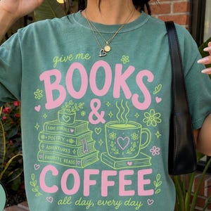 May include: A light green t-shirt featuring the pink and light green text "give me BOOKS & COFFEE all day, every day". The graphic includes a stack of books labeled "love stories", "poetry", "adventures", and "favorite reads" with a coffee mug.