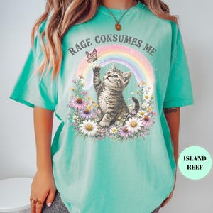 May include: A turquoise t-shirt featuring a graphic of a kitten reaching for a butterfly in front of a rainbow. The words "RAGE CONSUMES ME" are arched above the rainbow. The kitten is surrounded by flowers.