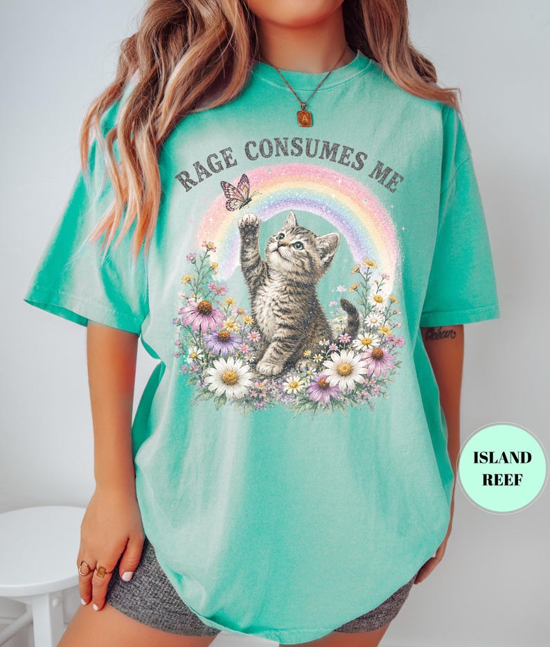 May include: A turquoise t-shirt featuring a graphic of a kitten reaching for a butterfly in front of a rainbow. The words "RAGE CONSUMES ME" are arched above the rainbow. The kitten is surrounded by flowers.