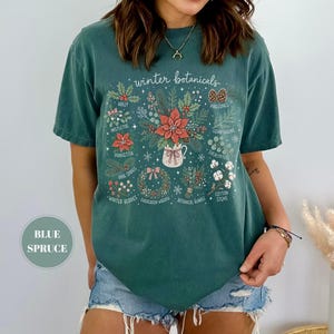 May include: A blue spruce colored t-shirt featuring a graphic print titled winter botanicals. The design displays various holiday plants including holly, mistletoe, poinsettia, pine sprigs, winter berries, pinecones, cedar greens, eucalyptus, and cotton stems.