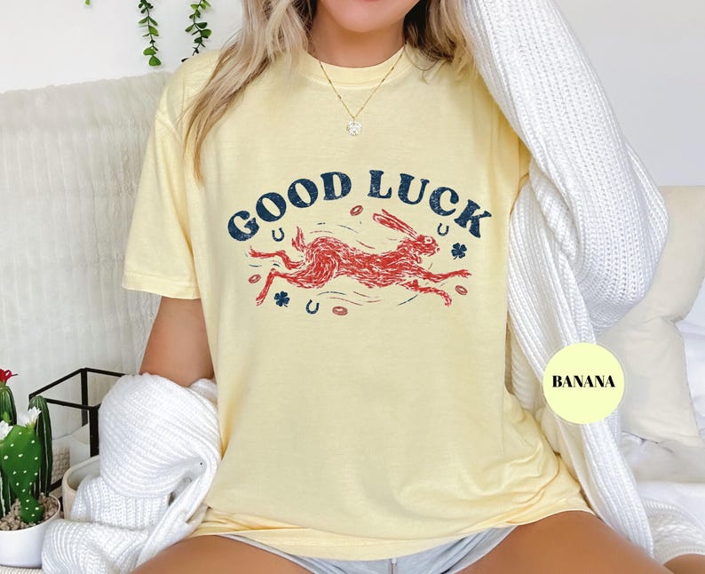 K&ouml;nnte beinhalten: Hellgelbes T-Shirt mit dem Schriftzug "GOOD LUCK" in marineblauer Schrift &uuml;ber einer roten Hasenillustration. Das Shirt zeigt auch Hufeisen und Kleebl&auml;tter.
