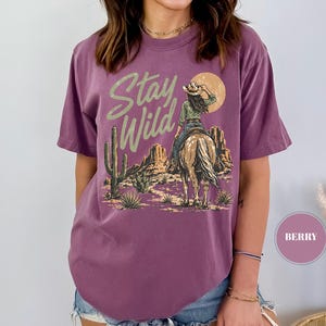 May include: Berry-coloured t-shirt featuring a desert scene with a cowgirl on horseback, cacti, and mountains. The text "Stay Wild" is printed above the image in a stylized font. The design includes a setting sun.