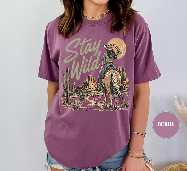 May include: Berry-coloured t-shirt featuring a desert scene with a cowgirl on horseback, cacti, and mountains. The text "Stay Wild" is printed above the image in a stylized font. The design includes a setting sun.