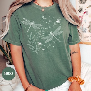 May include: A moss green t-shirt featuring a white illustration of dragonflies, bees, and dandelions. The design includes a crescent moon and hearts. The shirt has short sleeves and a crew neck.