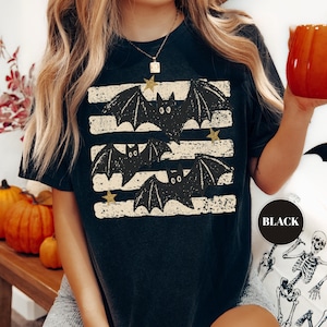 May include: A black short-sleeved t-shirt featuring a graphic print of three black bats flying over horizontal distressed white stripes, accented with small gold stars. The shirt has a round neckline and a relaxed fit, with the word BLACK printed.