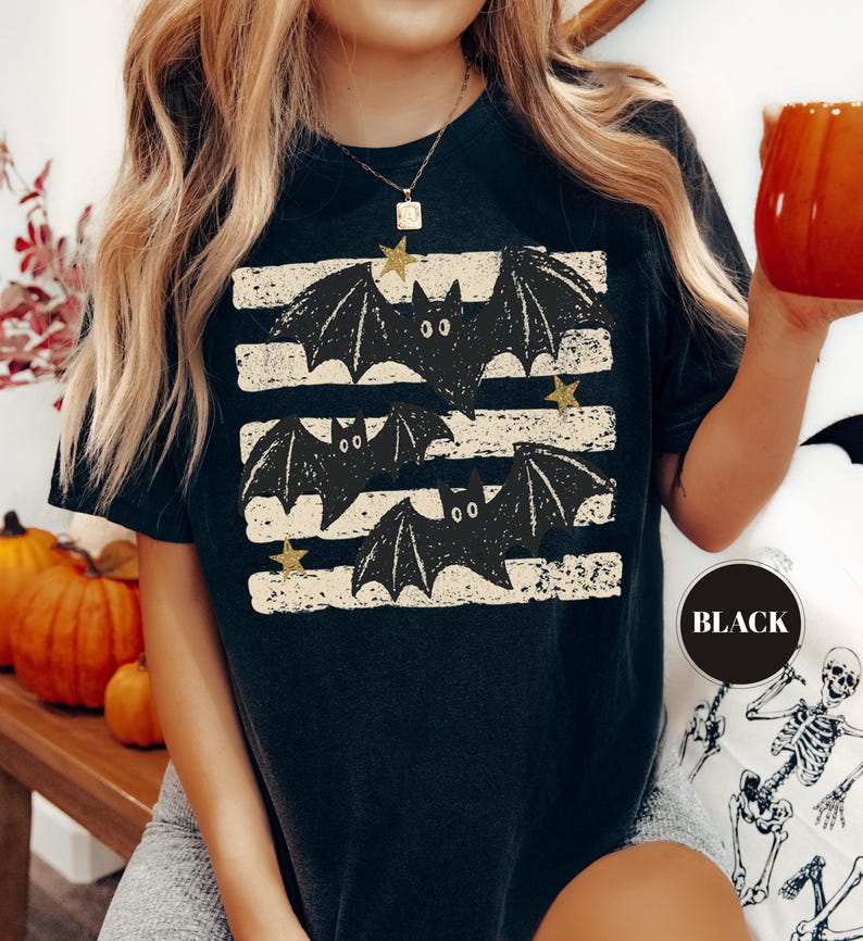 May include: A black short-sleeved t-shirt featuring a graphic print of three black bats flying over horizontal distressed white stripes, accented with small gold stars. The shirt has a round neckline and a relaxed fit, with the word BLACK printed.