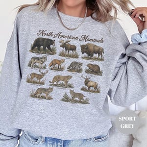 May include: A light grey crewneck sweatshirt featuring a graphic print titled North American Mammals. The design displays detailed illustrations of various animals including a black bear, moose, bison, wolf, fox, raccoon, beaver, deer, cougar, elk, coyote, and bobcat.