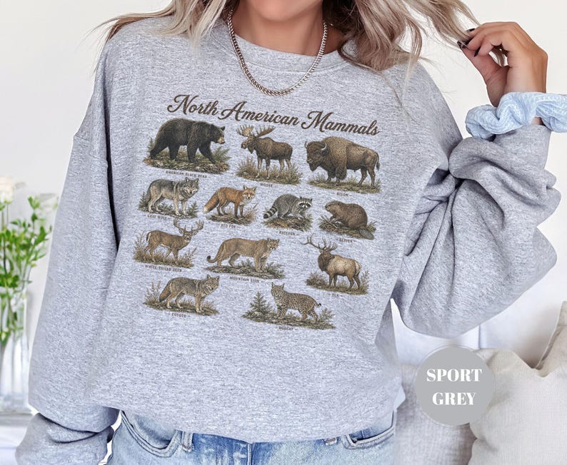 May include: A light grey crewneck sweatshirt featuring a graphic print titled North American Mammals. The design displays detailed illustrations of various animals including a black bear, moose, bison, wolf, fox, raccoon, beaver, deer, cougar, elk, coyote, and bobcat.