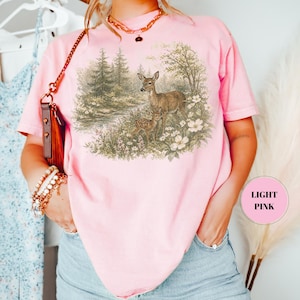 May include: A light pink t-shirt featuring a detailed illustration of two deer in a forest setting. The artwork includes trees, flowers, and a small stream. The text "LIGHT PINK" is in a pink circle on the right side of the shirt.