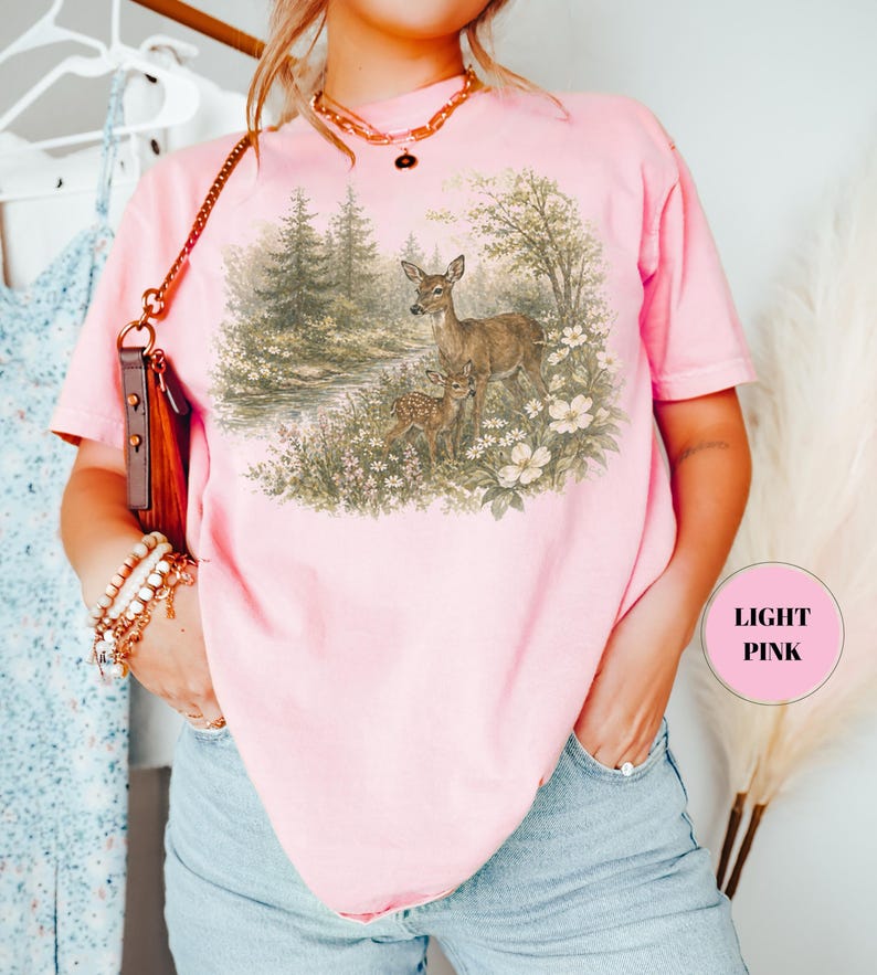 May include: A light pink t-shirt featuring a detailed illustration of two deer in a forest setting. The artwork includes trees, flowers, and a small stream. The text "LIGHT PINK" is in a pink circle on the right side of the shirt.
