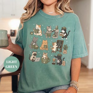May include: Light green t-shirt featuring a whimsical design of various cats engaged in coffee-related activities. The cats are depicted with coffee cups, brewing equipment, and reading books. The shirt has the words "Coffee First" and "Coffee Makes Everything Purr-fect".