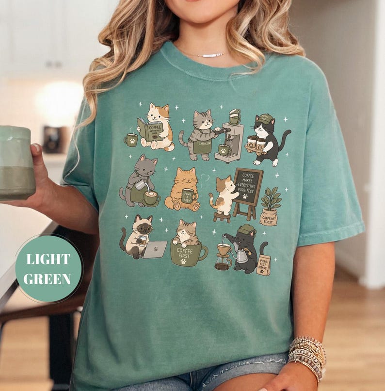 May include: Light green t-shirt featuring a whimsical design of various cats engaged in coffee-related activities. The cats are depicted with coffee cups, brewing equipment, and reading books. The shirt has the words "Coffee First" and "Coffee Makes Everything Purr-fect".