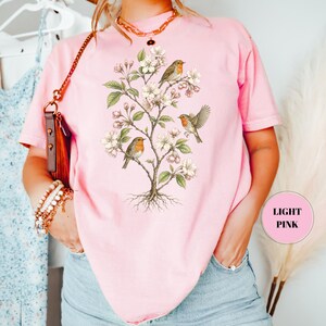 May include: Light pink t-shirt featuring a botanical illustration of a flowering tree with three robins. The birds have orange breasts and are perched on branches with white blossoms and green leaves. The shirt is a soft, solid color.
