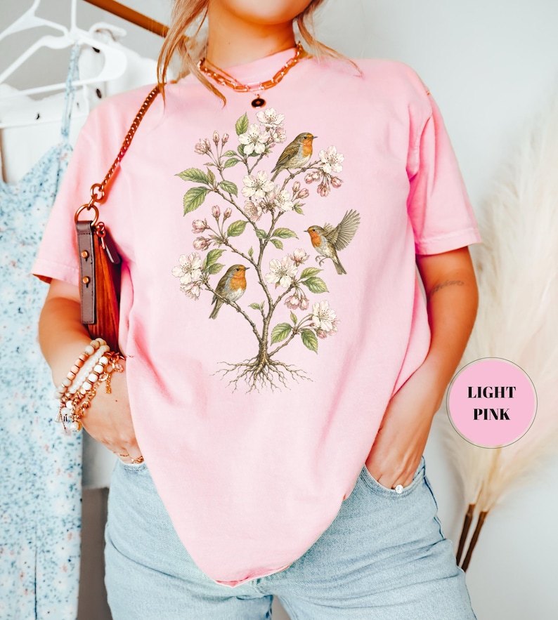 May include: Light pink t-shirt featuring a botanical illustration of a flowering tree with three robins. The birds have orange breasts and are perched on branches with white blossoms and green leaves. The shirt is a soft, solid color.