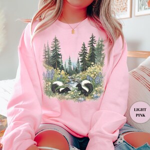 May include: Light pink sweatshirt featuring a nature scene with two skunks, a stream, and a forest. The graphic is detailed with wildflowers and trees. The sweatshirt is a solid light pink color.