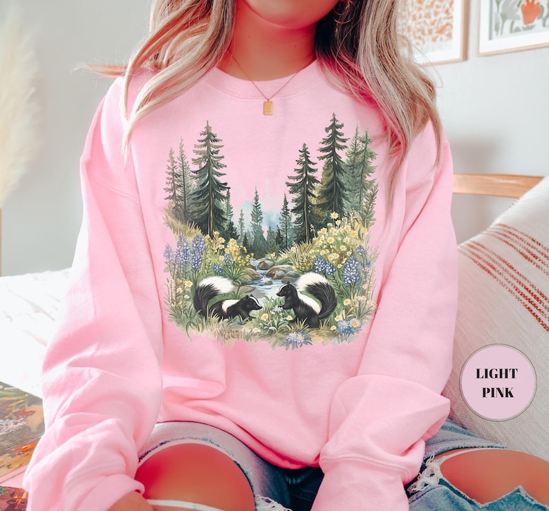 May include: Light pink sweatshirt featuring a nature scene with two skunks, a stream, and a forest. The graphic is detailed with wildflowers and trees. The sweatshirt is a solid light pink color.