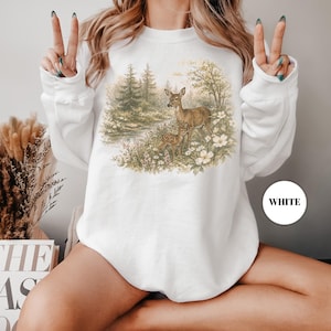 May include: White sweatshirt featuring a detailed illustration of deer in a forest scene. The artwork includes a doe and two fawns amidst trees, flowers, and a small stream. The word "WHITE" is printed on a white circle.