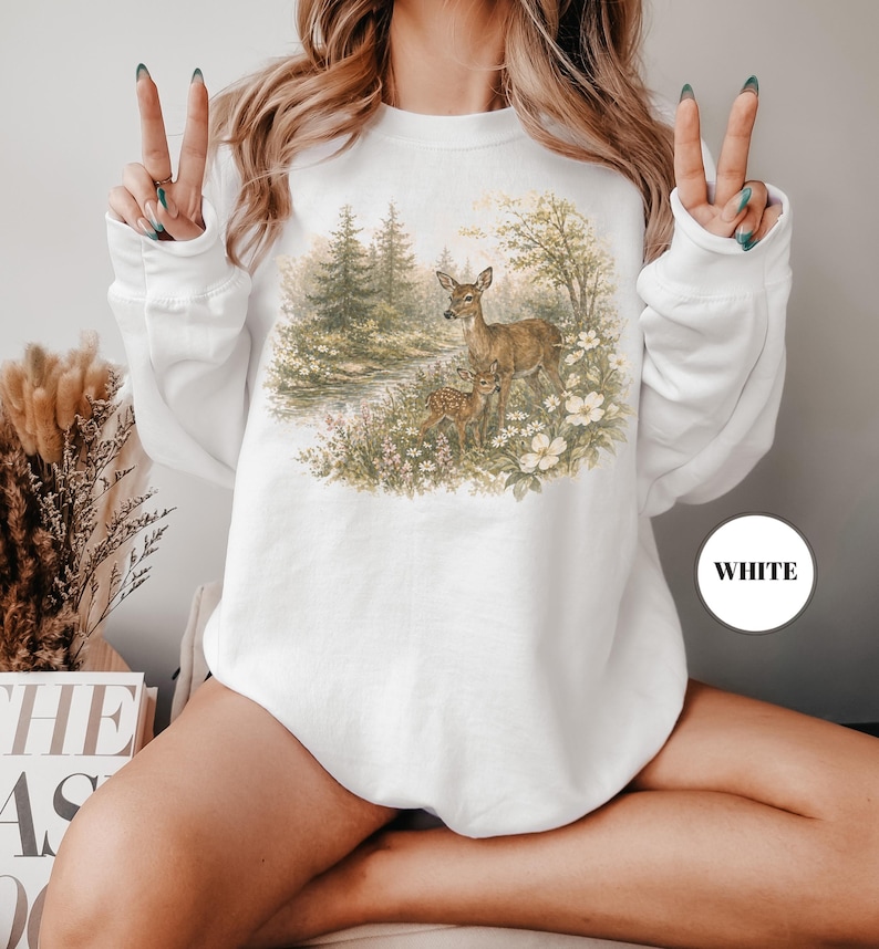 May include: White sweatshirt featuring a detailed illustration of deer in a forest scene. The artwork includes a doe and two fawns amidst trees, flowers, and a small stream. The word "WHITE" is printed on a white circle.