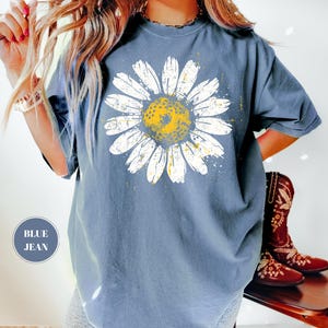 Puede incluir: Camiseta azul vaquera con un gran gr&aacute;fico de margarita. La margarita tiene p&eacute;talos blancos y un centro amarillo. La camiseta tiene un corte holgado y mangas cortas. El texto "BLUE JEAN" est&aacute; en un c&iacute;rculo en la parte inferior izquierda.