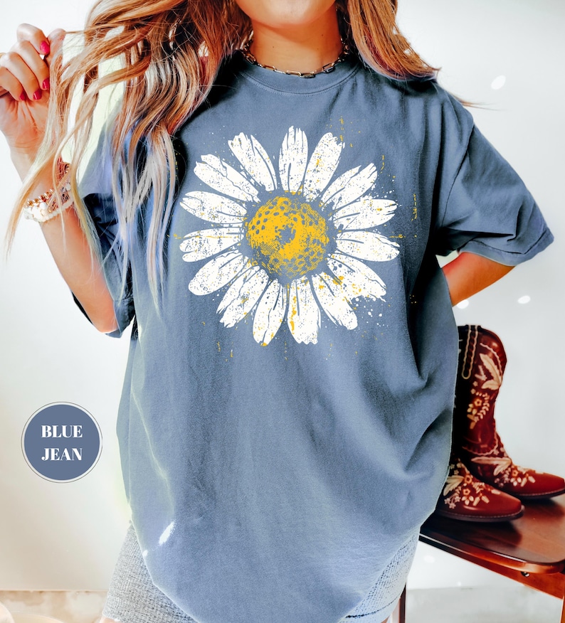 Puede incluir: Camiseta azul vaquera con un gran gr&aacute;fico de margarita. La margarita tiene p&eacute;talos blancos y un centro amarillo. La camiseta tiene un corte holgado y mangas cortas. El texto "BLUE JEAN" est&aacute; en un c&iacute;rculo en la parte inferior izquierda.