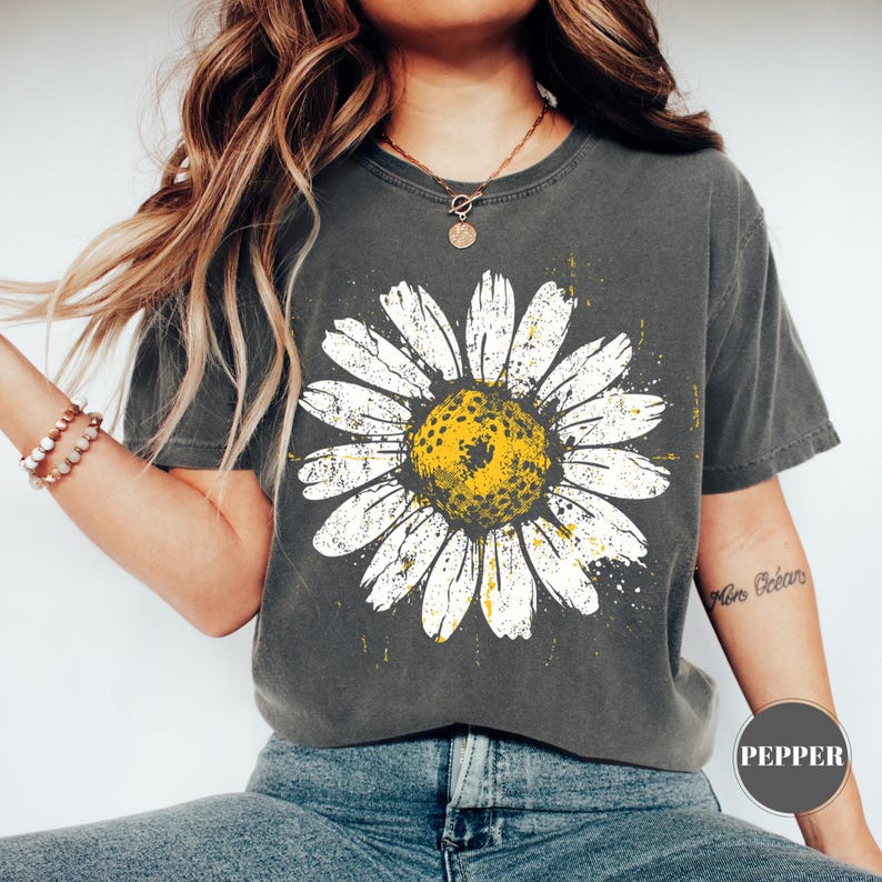 Puede incluir: Camiseta gris oscuro con una gran margarita blanca y centro amarillo. El dise&ntilde;o de la margarita tiene un aspecto envejecido y vintage. La camiseta es de cuello redondo y tiene mangas cortas. La palabra "Pepper" est&aacute; impresa en la parte inferior derecha.
