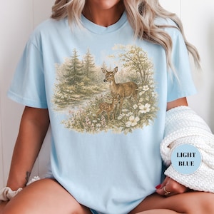 May include: Light blue t-shirt featuring a nature scene with two deer in a forest setting. The graphic includes trees, flowers, and a small body of water. The text "LIGHT BLUE" is in a circle on the shirt.