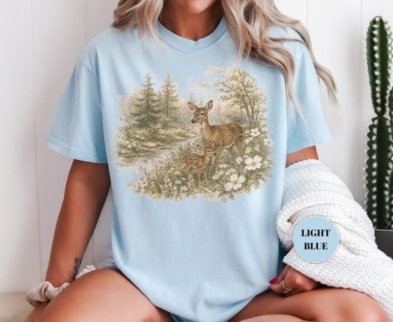 May include: Light blue t-shirt featuring a nature scene with two deer in a forest setting. The graphic includes trees, flowers, and a small body of water. The text "LIGHT BLUE" is in a circle on the shirt.