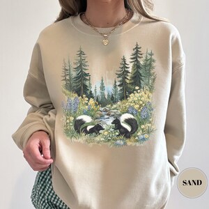 May include: Sand-colored sweatshirt featuring a nature scene with two skunks, a stream, and a forest. The graphic includes blue and yellow wildflowers and tall green trees. The word "SAND" is printed on a circular label in the lower right corner.