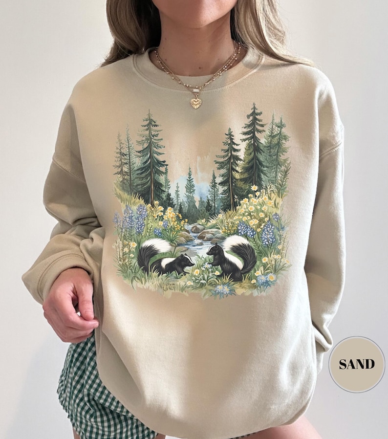 May include: Sand-colored sweatshirt featuring a nature scene with two skunks, a stream, and a forest. The graphic includes blue and yellow wildflowers and tall green trees. The word "SAND" is printed on a circular label in the lower right corner.