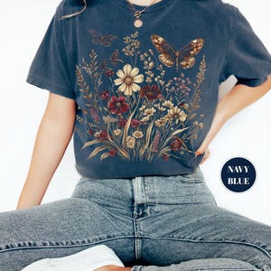 May include: A navy blue short-sleeve t-shirt featuring a graphic print of a wildflower bouquet with various blossoms in shades of cream, red, and brown, accompanied by two butterflies. The shirt has a round neckline and a relaxed fit.