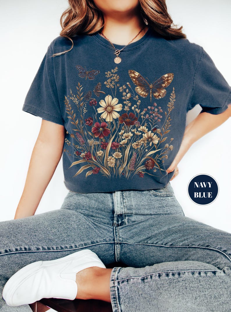 May include: A navy blue short-sleeve t-shirt featuring a graphic print of a wildflower bouquet with various blossoms in shades of cream, red, and brown, accompanied by two butterflies. The shirt has a round neckline and a relaxed fit.