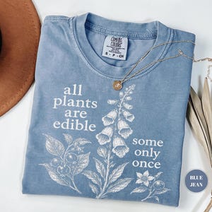 以下が含まれることがあります： all plants are edible some only onceというテキストと植物のイラストが白でプリントされた、ブルーデニムカラーのTシャツ。クルーネックで、襟の内側にComfort Colorsのブランドラベルが付いています。