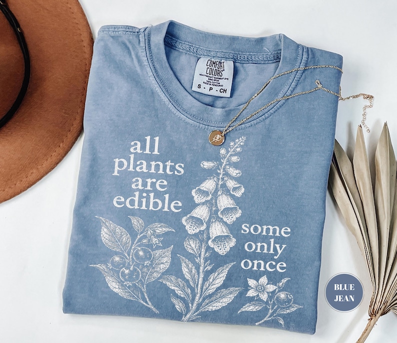 以下が含まれることがあります： all plants are edible some only onceというテキストと植物のイラストが白でプリントされた、ブルーデニムカラーのTシャツ。クルーネックで、襟の内側にComfort Colorsのブランドラベルが付いています。