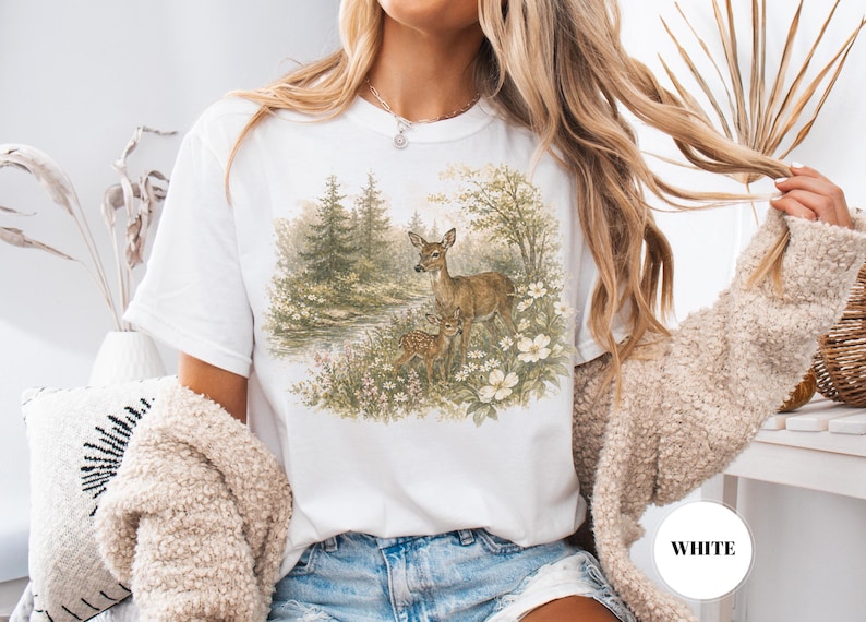 May include: White t-shirt featuring a watercolor illustration of two deer in a forest scene with trees, flowers, and a stream. The shirt has a classic crew neck and short sleeves. The word "WHITE" is in a white circle.
