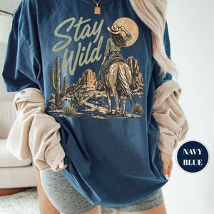 May include: Navy blue t-shirt with a desert-themed graphic. The design features a cowgirl on horseback, cacti, and the words "Stay Wild" in a stylized font. The shirt is a relaxed fit.