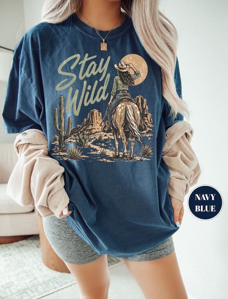 May include: Navy blue t-shirt with a desert-themed graphic. The design features a cowgirl on horseback, cacti, and the words "Stay Wild" in a stylized font. The shirt is a relaxed fit.