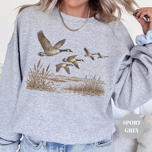 Peut inclure: Un sweat-shirt &agrave; col rond gris sport orn&eacute; d'une illustration d&eacute;taill&eacute;e repr&eacute;sentant un vol d'oies du Canada au-dessus d'un paysage mar&eacute;cageux avec des roseaux. Le motif utilise des tons naturels de brun et de beige pour les oiseaux et le d&eacute;cor.