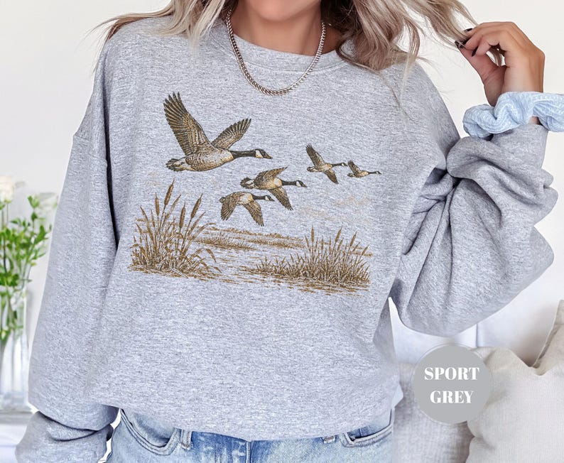 Peut inclure: Un sweat-shirt &agrave; col rond gris sport orn&eacute; d'une illustration d&eacute;taill&eacute;e repr&eacute;sentant un vol d'oies du Canada au-dessus d'un paysage mar&eacute;cageux avec des roseaux. Le motif utilise des tons naturels de brun et de beige pour les oiseaux et le d&eacute;cor.