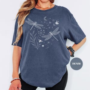 May include: A denim blue t-shirt featuring a white illustration of two dragonflies, a crescent moon, stars, and bees. The design includes floral elements and dandelion seeds. The shirt is a relaxed fit.