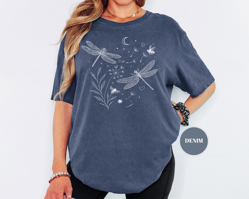 May include: A denim blue t-shirt featuring a white illustration of two dragonflies, a crescent moon, stars, and bees. The design includes floral elements and dandelion seeds. The shirt is a relaxed fit.