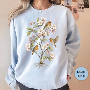 May include: Light blue sweatshirt featuring a detailed illustration of a flowering tree with three robins. The birds have orange breasts and are perched on branches with white and pink blossoms. The sweatshirt has a round neckline and long sleeves. The text "LIGHT BLUE" is in a light blue circle.
