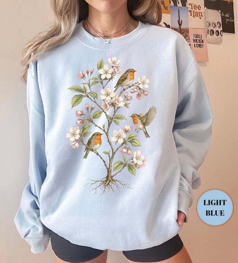 May include: Light blue sweatshirt featuring a detailed illustration of a flowering tree with three robins. The birds have orange breasts and are perched on branches with white and pink blossoms. The sweatshirt has a round neckline and long sleeves. The text "LIGHT BLUE" is in a light blue circle.