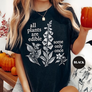 以下が含まれることがあります： 黒いTシャツで、フォックスグローブの植物とベリーの白い植物画が描かれています。胸の中央には「all plants are edible some only once」というテキストが、はっきりとした白いセリフ体でプリントされています。