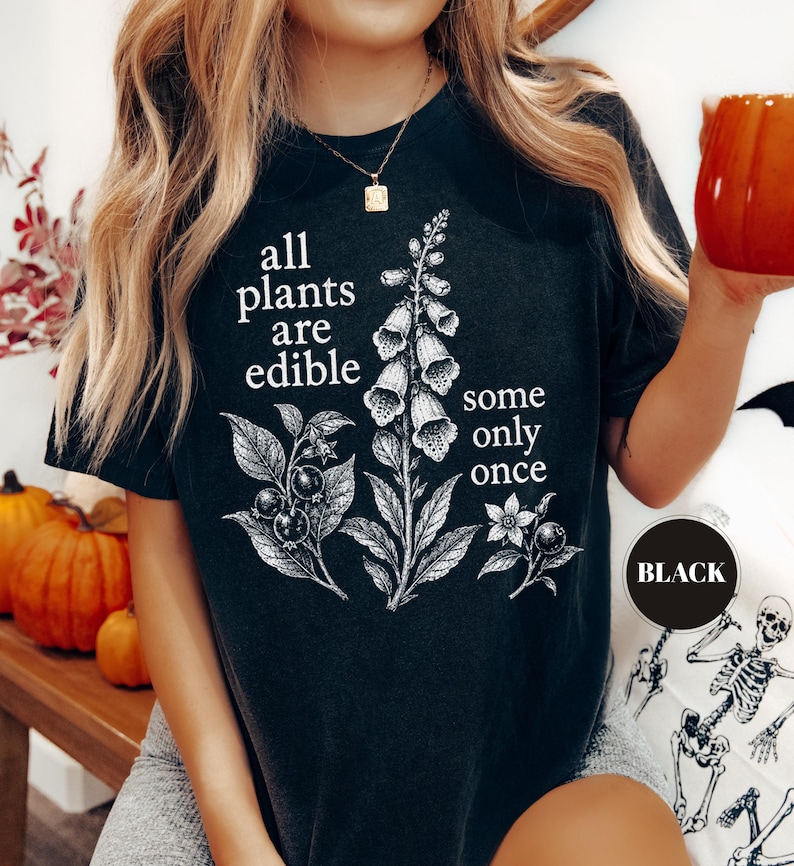 以下が含まれることがあります： 黒いTシャツで、フォックスグローブの植物とベリーの白い植物画が描かれています。胸の中央には「all plants are edible some only once」というテキストが、はっきりとした白いセリフ体でプリントされています。