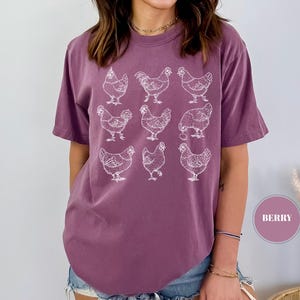 May include: Berry-colored t-shirt featuring a white graphic of nine chickens. The short-sleeved shirt has a relaxed fit. The design is a simple line drawing of chickens in various poses.