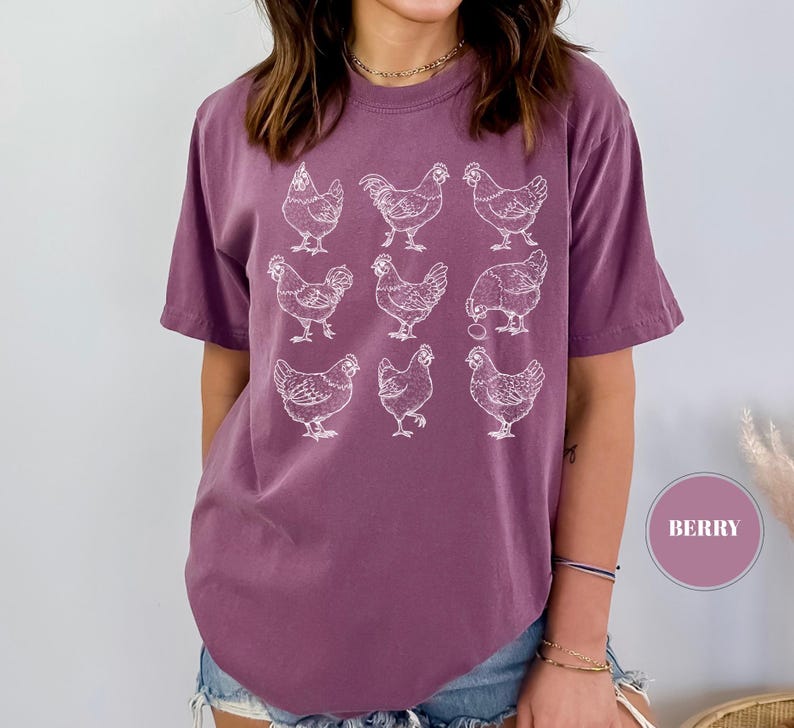 May include: Berry-colored t-shirt featuring a white graphic of nine chickens. The short-sleeved shirt has a relaxed fit. The design is a simple line drawing of chickens in various poses.