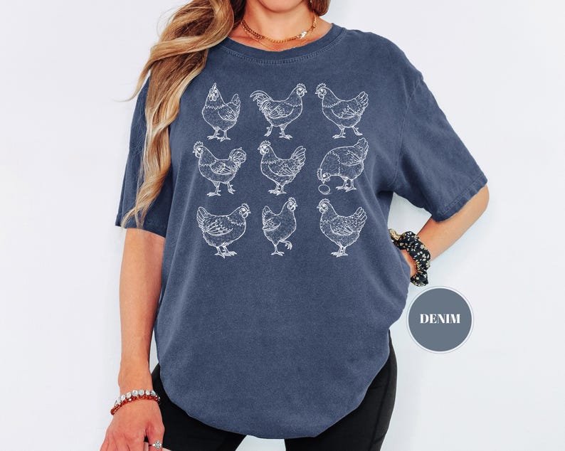 May include: A denim blue t-shirt featuring a white illustration of nine chickens in various poses. The short-sleeved shirt has a crew neck and a relaxed fit. The word "DENIM" is in a gray circle.