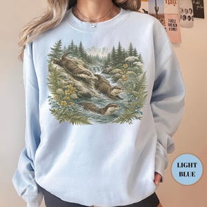Puede incluir: Una sudadera azul claro de cuello redondo con un estampado gr&aacute;fico detallado de tres nutrias nadando en un arroyo de monta&ntilde;a. La ilustraci&oacute;n incluye pinos verdes, helechos, flores silvestres amarillas y una cascada ante monta&ntilde;as.
