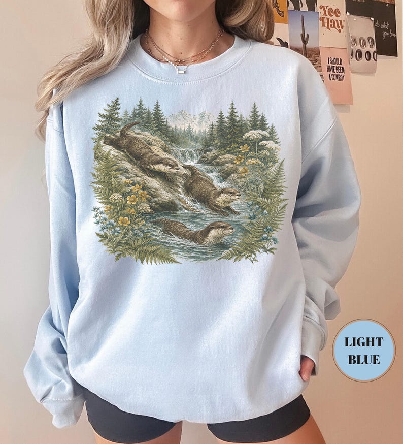 Puede incluir: Una sudadera azul claro de cuello redondo con un estampado gr&aacute;fico detallado de tres nutrias nadando en un arroyo de monta&ntilde;a. La ilustraci&oacute;n incluye pinos verdes, helechos, flores silvestres amarillas y una cascada ante monta&ntilde;as.